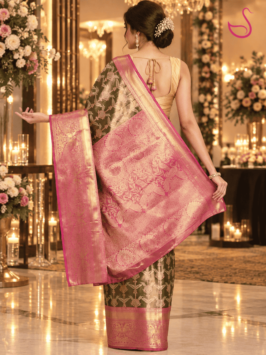 Woman shown from the back wearing a dark olive-green and pink-gold brocade saree, highlighting the richly woven pink-gold pallu and blouse back, posed in a luxurious reception setting with warm lighting, floral décor and candles. Ishaana Ethnics emblem appears in the top-right corner.