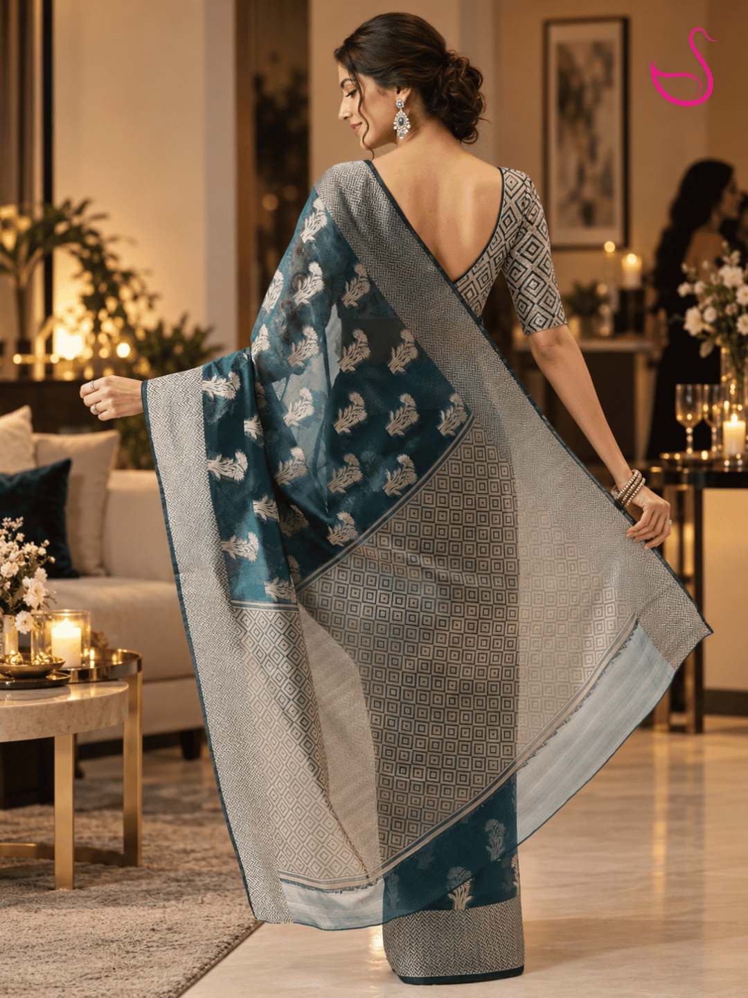 woman shown from the back wearing a teal-blue saree with intricate silver floral motifs, geometric woven border and detailed pallu, paired with a geometric-patterned V-back blouse and traditional jewellery, photographed in a warm, elegant house-party setting. Pink swan emblem appears at the top right.