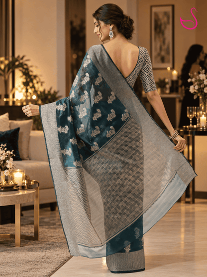 woman shown from the back wearing a teal-blue saree with intricate silver floral motifs, geometric woven border and detailed pallu, paired with a geometric-patterned V-back blouse and traditional jewellery, photographed in a warm, elegant house-party setting. Pink swan emblem appears at the top right.