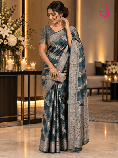 Model wearing a deep teal blue Banarasi saree with intricate silver floral zari motifs, ornate silver zari border and detailed woven pallu, paired with a statement blouse and jewellery.
