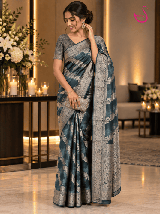 Model wearing a deep teal blue Banarasi saree with intricate silver floral zari motifs, ornate silver zari border and detailed woven pallu, paired with a statement blouse and jewellery.