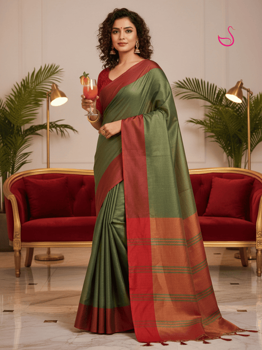 A woman models a green and maroon saree in a decorated indoor setting with maroon sofas and plants. The saree has a sage green body with a wide maroon border and pallu.