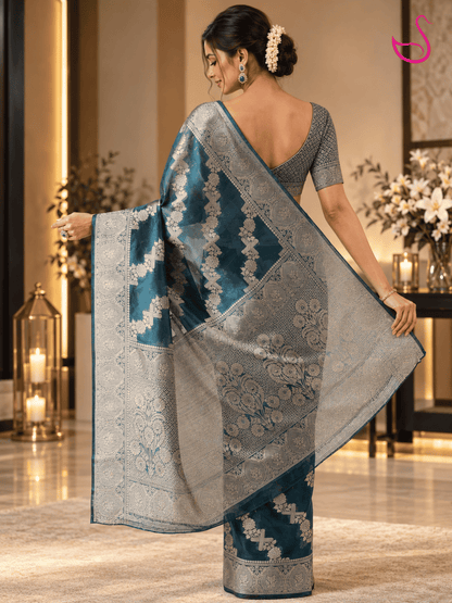 Back view of a model wearing a deep teal blue Banarasi saree showcasing its intricate silver floral weave, detailed zari border and richly patterned pallu, paired with a statement blouse.