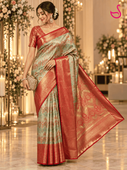 A woman models a traditional Indian saree in a decorated indoor event space with floral arrangements and candles. The saree has a light sage green body with intricate gold zari patterns and a rich coral-red border and pallu.