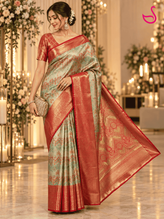 A woman models a traditional Indian saree in a decorated indoor event space with floral arrangements and candles. The saree has a light sage green body with intricate gold zari patterns and a rich coral-red border and pallu.