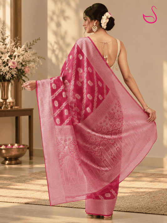 woman shown from the back in a vivid pink saree, displaying the intricate floral motifs, woven borders and richly patterned pallu, with a cream open-back blouse and traditional jewellery in a warm festive setting.