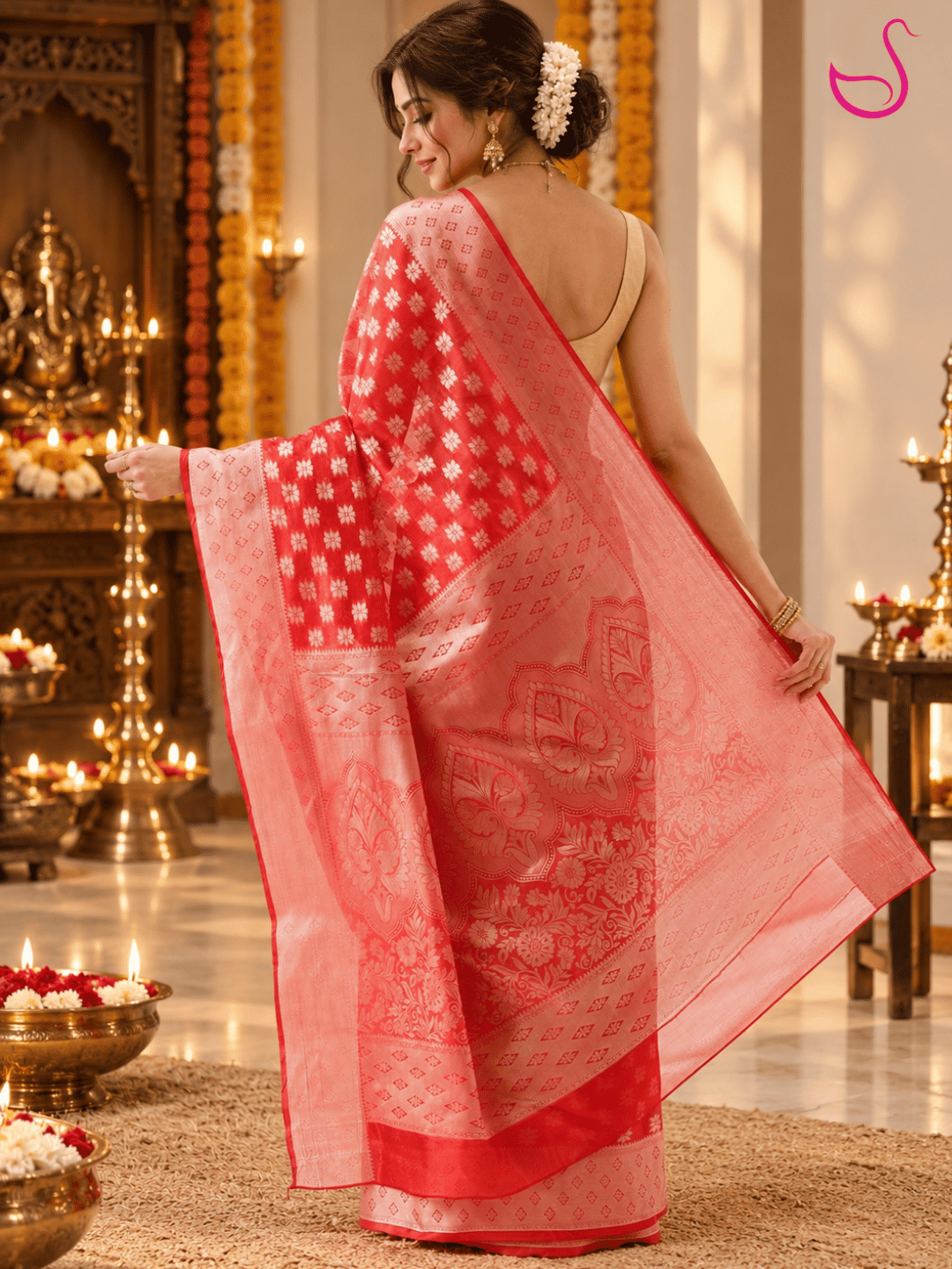 Woman wearing a red and peach-pink Banarasi saree with intricate floral motifs, ornate woven borders and a detailed pallu, styled with traditional jewellery and jasmine flowers, standing in a warmly lit festive pooja setting with brass lamps and floral decorations, with the Ishaana Ethnics emblem.