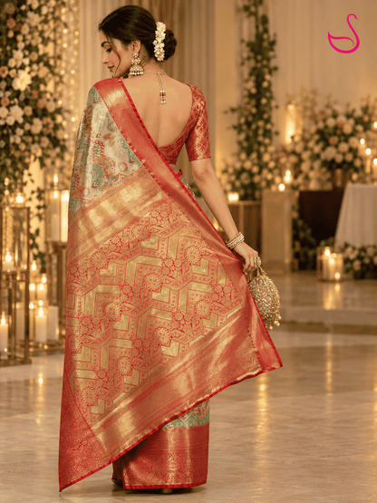 A woman wearing a traditional Indian saree, photographed from behind in an elegant indoor setting with warm lights and floral decorations. The saree has a red border, a gold and red patterned body, and a light green and gold border.
