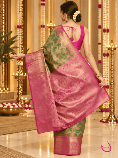 A woman wearing a traditional Indian saree, photographed from behind in an ornate indoor setting. The saree has a vibrant pink border and blouse, with a green body featuring gold floral patterns and a matching pink and gold border.