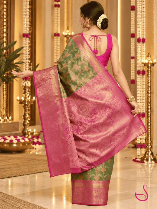 A woman wearing a traditional Indian saree, photographed from behind in an ornate indoor setting. The saree has a vibrant pink border and blouse, with a green body featuring gold floral patterns and a matching pink and gold border.