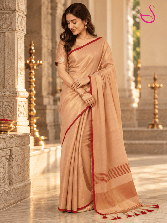 A woman wearing a beige silk saree with red border accents, standing in a traditional stone corridor with brass lamps.