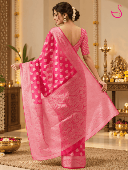 Back view of a woman wearing a vibrant hot-pink Banarasi saree featuring intricate paisley motifs, ornate floral borders and detailed woven pallu, paired with a matching patterned blouse. She stands in a softly lit pooja setting with brass lamps, flowers and traditional décor. Pink swan emblem visible in the upper-right corner.