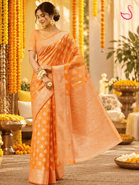 Woman wearing a vibrant orange woven saree with intricate gold motifs, detailed borders and ornate pallu, paired with a matching patterned blouse and floral jewellery, posed at a traditional Haldi ceremony.