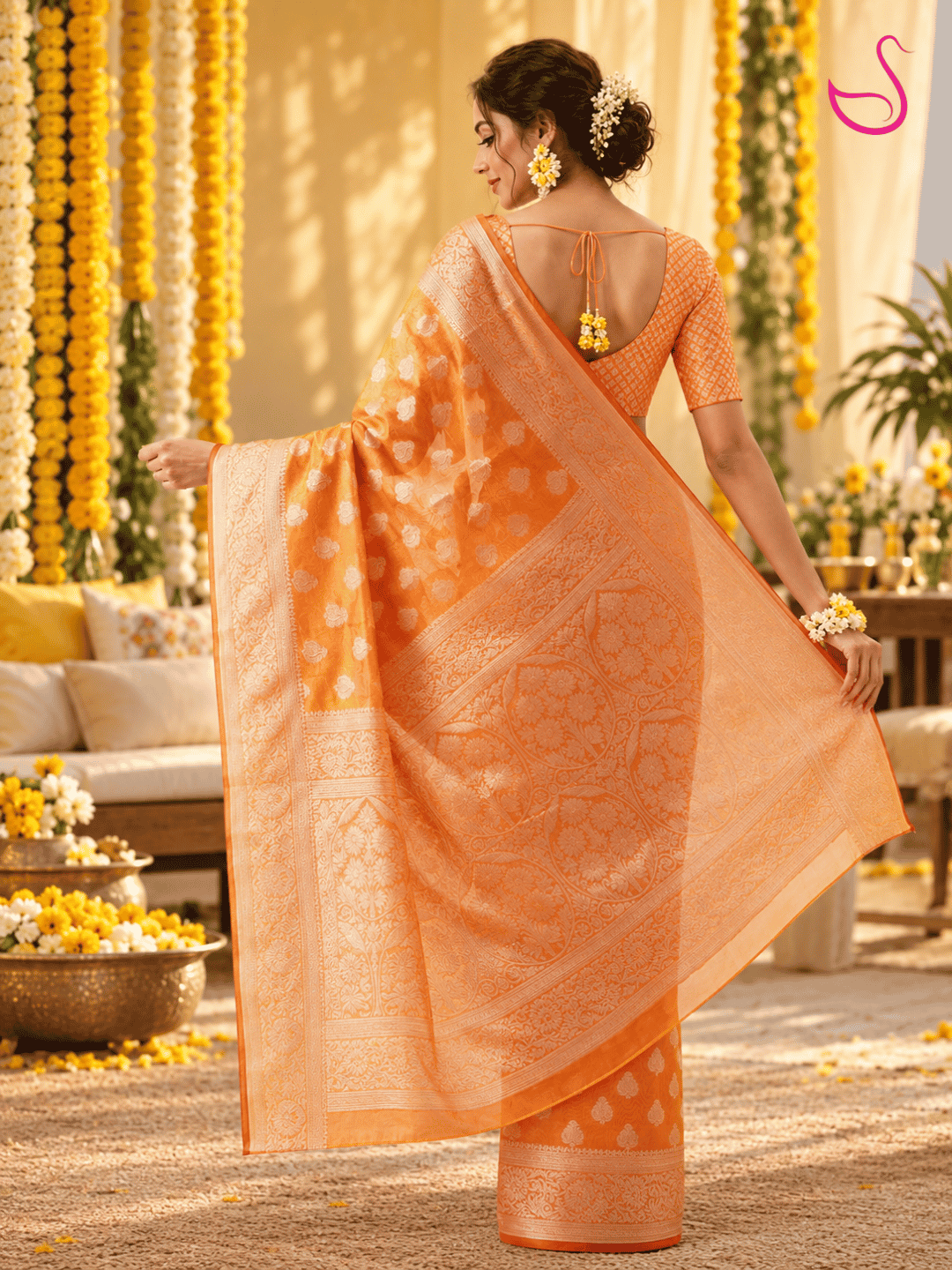 Woman shown from the back wearing the same vibrant orange woven saree with intricate gold motifs, detailed borders and ornate pallu, paired with a matching patterned blouse and floral jewellery, against a warm Haldi ceremony setting.