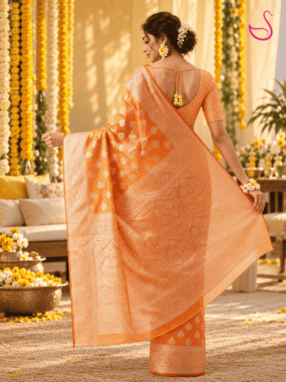 Woman shown from the back wearing the same vibrant orange woven saree with intricate gold motifs, detailed borders and ornate pallu, paired with a matching patterned blouse and floral jewellery, against a warm Haldi ceremony setting.