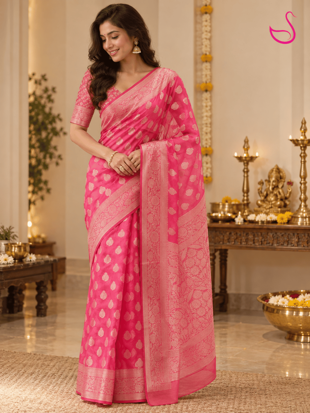 Woman wearing a vibrant hot-pink Banarasi saree with intricate woven paisley motifs, floral borders and detailed pallu, paired with a matching patterned blouse, posed in an elegant pooja setting with brass lamps, flowers and traditional décor. Pink swan emblem visible in the upper-right corner.
