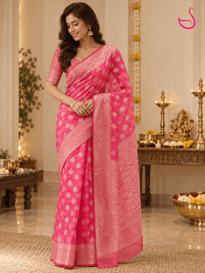 Woman wearing a vibrant hot-pink Banarasi saree with intricate woven paisley motifs, floral borders and detailed pallu, paired with a matching patterned blouse, posed in an elegant pooja setting with brass lamps, flowers and traditional décor. Pink swan emblem visible in the upper-right corner.