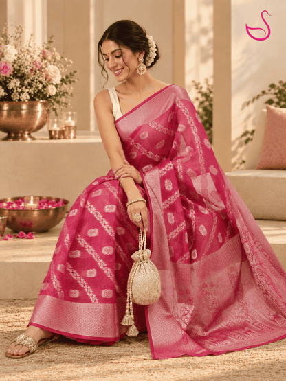 woman seated gracefully in a vivid pink saree with delicate pale floral motifs, ornate woven borders and a detailed patterned pallu, styled with traditional jewellery and a cream blouse in an elegant festive setting.