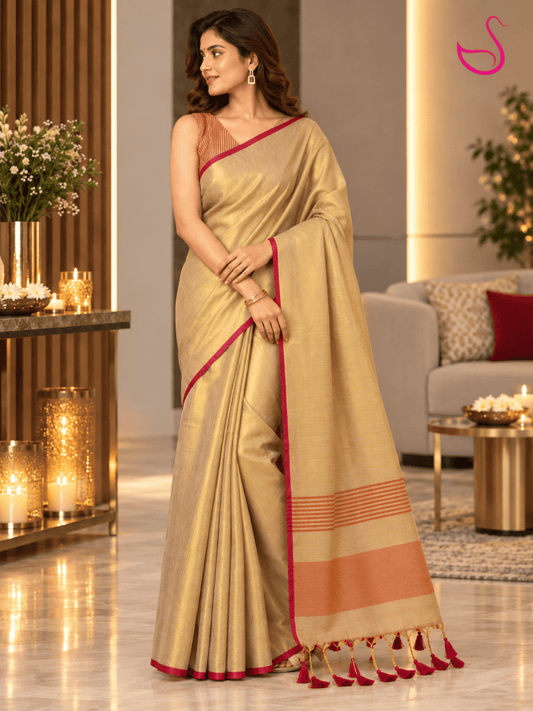 A woman models a beige silk saree with red and orange border accents in a modern, warmly lit living room setting.