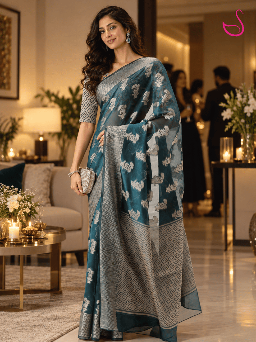 woman wearing a teal-blue saree with intricate silver floral motifs, geometric woven border and detailed pallu, styled with a patterned blouse and traditional jewellery, photographed in a warm, elegant house-party setting. Pink swan emblem appears at the top right.