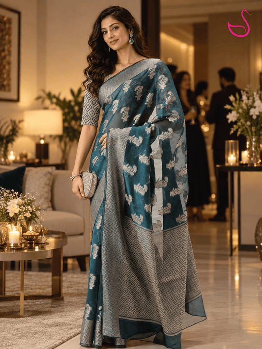 woman wearing a teal-blue saree with intricate silver floral motifs, geometric woven border and detailed pallu, styled with a patterned blouse and traditional jewellery, photographed in a warm, elegant house-party setting. Pink swan emblem appears at the top right.