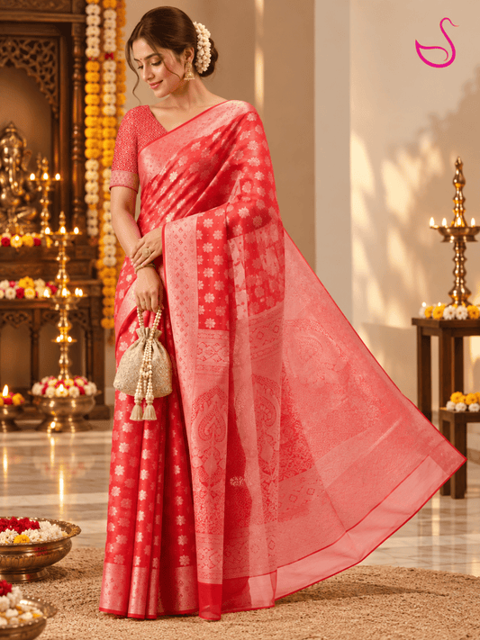 Woman wearing a red and peach-pink Banarasi saree with intricate floral motifs, traditional woven borders and a detailed pallu, posed against a dark elegant interior with a vase of flowers and the Ishaana Ethnics emblem.