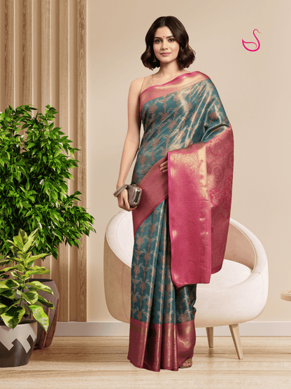 A woman models a teal and magenta saree with gold zari detailing in a styled indoor setting with a white chair and potted plants.