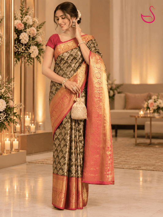 woman wearing an olive-green silk saree with intricate gold floral motifs, a rich gold zari border and vibrant red-orange pallu, styled with traditional jewellery and a decorative potli bag in an elegant indoor setting.